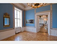 Eleganter Innenraum eines historischen Gebäudes mit blauen Wänden, verziertem weißen Holzpaneling und Parkettboden. Rechts führt eine Tür in einen Saal mit Kronleuchtern, weißen Stühlen und hohen Fenstern.