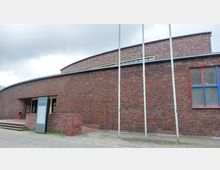 Backsteingebäude mit abgerundeter Fassade, einem Eingangsbereich links und einem Schild mit der Aufschrift "Max-Taut-Aula". Rechts befinden sich große Fenster mit auffällig blau-roten Rahmen und einige Fahnenmasten vor der Fassade.