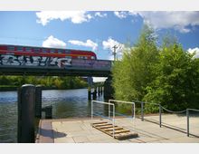 Eine Fußgängerplattform mit Geländern am Ufer eines Flusses, im Hintergrund eine Brücke mit einer vorbeifahrenden roten Regionalbahn und Graffiti. Auf der rechten Seite dichter grüner Bewuchs und blauer Himmel mit Wolken.