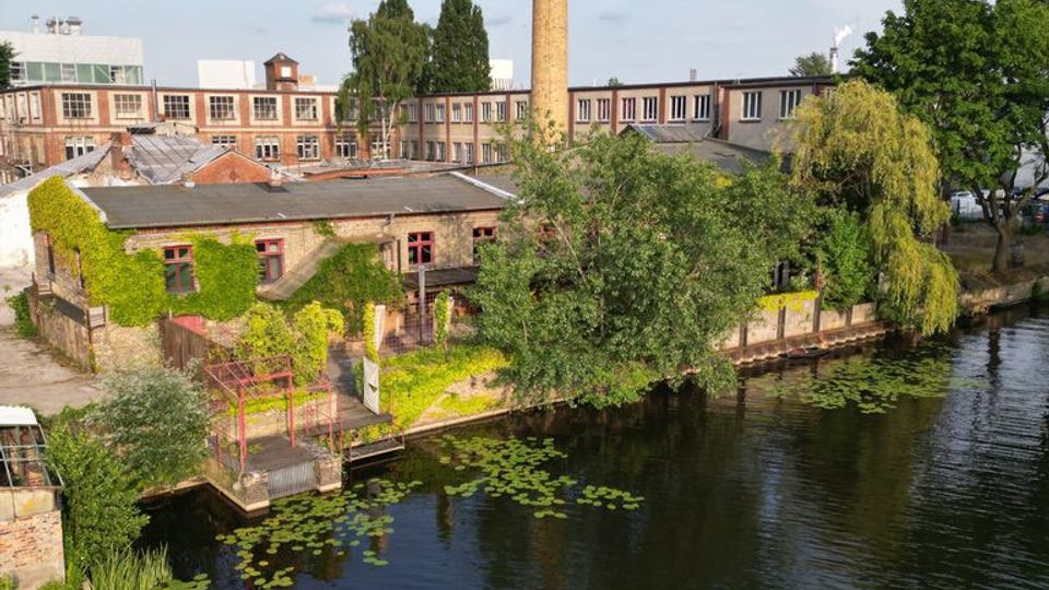 Blick über einen Kanal mit Seerosen auf ein ehemaliges Industrieareal mit Backsteingebäuden, bewachsenen Fassaden und einem hohen Schornstein. Am Ufer stehen Bäume, und im Wasser liegt eine gelb-weiße Boje.