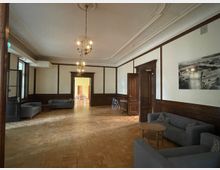 Ein historisch anmutender Raum mit Holzvertäfelungen an den Wänden und einem Parkettboden. Der Raum enthält mehrere graue Sofas, einen kleinen Holztisch und dekorative Kronleuchter an der Decke; im Hintergrund ist ein Durchgang zu einem weiteren Raum mit Holzmöbeln sichtbar.