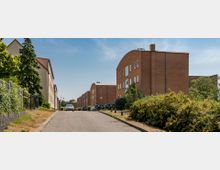 Straßenszene in einem Wohngebiet mit modernen Backstein-Wohngebäuden; links ein Gehweg mit Zaun und Bäumen, rechts Büsche und parkende Autos. Die Straße verläuft leicht bergab unter einem klaren, blauen Himmel.