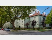 Mehrstöckiges Wohnhaus mit grün gestrichener Fassade und roten Ziegeldächern, umgeben von Bäumen und Büschen. Im Vordergrund sind parkende Autos, ein Gehweg und eine Straßenlaterne zu sehen.