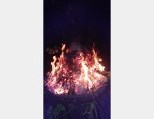 Ein brennendes Lagerfeuer in einer Feuerschale, umgeben von Zweigen und Blättern, mit Funken und Flammen, die in die Dunkelheit aufsteigen. Der Hintergrund ist dunkel und hebt das Feuer hervor.