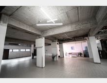 Offener, industrieller Raum mit Sichtbetondecke, weißen Säulen und gefliesten Böden. Im Hintergrund befindet sich eine minimalistisch eingerichtete Sitzbereich mit einem Sofa, einer Bank und einigen Pflanzen.