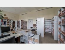 Büroarchivraum mit mehreren Regalen, die dicht mit Akten gefüllt sind, sowie einem Schreibtisch mit Monitor, Tastatur und Büromaterialien. Im Hintergrund ist eine Tür, und an den Wänden hängen ein Kalender und eine Uhr.