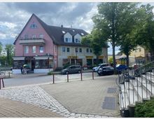 Straßenszene mit mehreren zweigeschossigen Gebäuden, darunter ein rosa Haus mit spitzem Dach und Balkonen, sowie Geschäften im Erdgeschoss. Im Vordergrund sind Pflastersteine, eine Treppe mit Geländer und parkende Autos entlang der Straße zu sehen, flankiert von Bäumen.