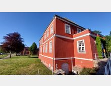 Zweistöckiges Gebäude in roter Farbe mit weißen Fensterrahmen und dekorativen weißen Fassadenlinien, umgeben von einer gepflegten Rasenfläche und Bäumen. Das Gebäude hat ein Satteldach und einen steinernen Sockel, mit einem kleinen Weg und einer mit Efeu bewachsenen Wand an der rechten Seite.