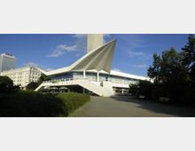 Modernes Gebäude mit futuristischer Dachkonstruktion und geschwungenen Formen, das auf Stützen erhöht steht und von einer breiten Treppe zugänglich ist. Im Hintergrund ist ein hoher Hotelturm sowie ein weiterer mehrstöckiger Gebäudekomplex sichtbar, umgeben von Bäumen und einer gepflasterten Fläche.