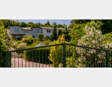 Mehrfamilienhaus in einer grünen, bewaldeten Umgebung, umgeben von dichtem Garten mit blühenden weißen Sträuchern und kleinen Bäumen; im Vordergrund ein grünes Metalltor und ein gepflasterter Weg.