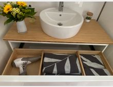 Weiße Waschbeckenschale auf einer Holzplatte mit einem kleinen Blumenstrauß links und einem Glas mit Deckel rechts. Darunter ein geöffneter Schubladeneinsatz mit Holzfächern, in denen ein Haartrockner und gefaltete Handtücher liegen.