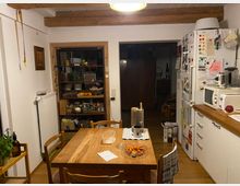 Eine Küche mit Holzbalkendecke und Holzboden. Im Vordergrund steht ein Esstisch aus Holz mit vier Stühlen, darauf befinden sich eine Karaffe, ein Glas, eine Tasse und ein Teller mit kleinen Tomaten. Rechts ist eine Küchenzeile mit weißer Front, auf der eine Mikrowelle und weitere Küchenutensilien stehen. Links sind ein Regal mit Vorräten und eine Heizung sichtbar.