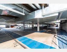 Ein modernes Parkhaus mit mehreren Parkreihen, markierten Parkplätzen und Säulen, die die Decke stützen. Die Decke besteht aus hellen Metallverkleidungen mit sichtbaren Lüftungsrohren, und die blau markierten Bereiche deuten auf spezielle Parkzonen hin.