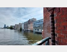 Blick auf eine Flusslandschaft mit modernen und historischen Backsteingebäuden am Ufer, darunter ein Gebäude mit großen Glasfassaden. Im Vordergrund ist ein Teil eines roten Backsteinbogens sichtbar.