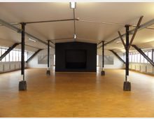 Großer, offener Raum mit Parkettboden, schwarzen Stahlstützen und einer schwarzen Wand mit quadratischer Vertiefung. Der Raum hat schräg verlaufende Dachbalken, große Fensterreihen an den Seiten und helle Deckenleuchten.