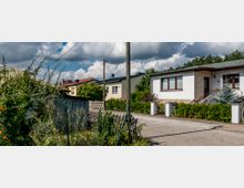 Eine Wohnsiedlung mit freistehenden Einfamilienhäusern, umgeben von gepflegten Gärten und Hecken. Im Vordergrund ist eine gepflasterte Straße sichtbar, daneben ein Zaun mit dichtem Pflanzenbewuchs.