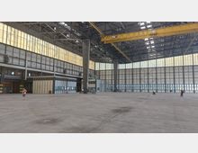 Große, leere Industriehalle mit hohen Decken, Stahlträgern und lichtdurchlässigen Wänden. Im Vordergrund eine offene Fläche, im Hintergrund kleinere Räume und Personen mit Warnwesten, sowie eine gelbe Laufkatze an der Decke.