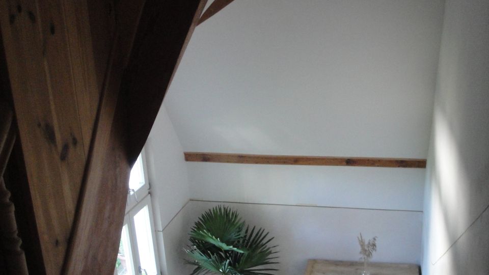 Treppenhaus mit Holztreppe und gedrechseltem Geländer unter einer hohen, schrägen Decke mit sichtbaren Holzbalken. Neben einem Fenster stehen eine Topfpflanze und eine niedrige Holzkommode auf grünem Teppichboden.