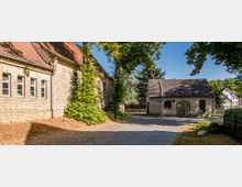 Eine gepflasterte Dorfstraße mit einem alten, aus gelben Ziegelsteinen gebauten Gebäude auf der linken Seite und einem kleinen, verputzten Haus mit Satteldach und grünem Holztor auf der rechten Seite. Im Hintergrund stehen Bäume und weitere Gebäude, die von hellem Sonnenlicht beleuchtet werden.