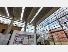 Großer moderner Innenraum mit Blick nach draußen durch eine hohe Glasfassade, die von Betonpfeilern gestützt wird. Draußen sind Bäume und ein historisches Backsteingebäude zu sehen, während im Vordergrund Poster und technische Ausstellungsstücke stehen.