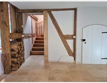 Innenraum eines Fachwerkhauses mit Holzbalken an den weißen Wänden. Im Vordergrund liegt ein Stapel Holzscheite, links führt eine Holztreppe nach oben, und rechts ist eine kleine weiße Tür mit einem schwarzen Türriegel sichtbar. Der Boden besteht aus beigefarbenen Fliesen.