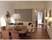 Wohnzimmer mit offener Raumaufteilung, einem Holztisch und vier Korbstühlen im Vordergrund. Im Hintergrund ein weißes Sofa vor einer schwarz-weiß gestreiften Wand, dekoriert mit Deckenlampen, Bildern, und Pflanzen; durch eine doppelflügelige Tür ist ein angrenzender Raum mit Kronleuchter sichtbar.