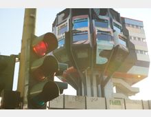 Ein moderner, futuristischer Gebäudeturm mit bunter Fassade und markantem Design, der auf Säulen ruht. Im Vordergrund ist eine Verkehrsampel mit rotem Licht zu sehen, während ein Zaun mit Graffiti das untere Viertel des Bildes einnimmt.