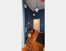 Flur in einem Altbau mit dunkelblauen Wänden und Holzfußboden. Im Vordergrund steht ein kleiner Schrank mit Dekoration, daneben Bücher und eine Lampe; von der Decke hängen mehrere Pendelleuchten. Eine Tür führt in einen angrenzenden, hell beleuchteten Raum.