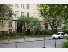 Mehrstöckiges Wohngebäude mit gelber Fassade, umgeben von Bäumen und einem kleinen Vorgarten mit Holzzaun. Im Vordergrund verläuft eine Straße mit einem geparkten weißen Auto und einem historischen Laternenpfosten.