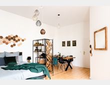Helles Schlafzimmer kombiniert mit einem Essbereich und skandinavischem Interieur-Design. Ein Bett mit grüner Decke, ein offenes Regal mit Pflanzen, ein Esstisch aus Holz mit Stühlen sowie dekorative Details wie Wandleisten und Hängelampen prägen den Raum.