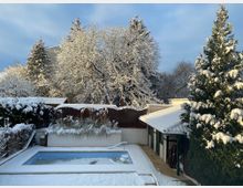 Verschneiter Garten mit leerem Whirlpool im Vordergrund, umgeben von Pflanzen und Bäumen, die mit Schnee bedeckt sind; im Hintergrund eine mit Schnee bedeckte Terrasse und ein kleines Gebäude mit Flachdach.