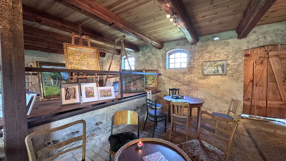 Rustikaler Innenraum mit freiliegenden Holzbalken, groben Steinwänden und kleinen Rundbogenfenstern. Eine erhöhte Plattform zeigt gerahmte Gemälde und Kunstwerke, davor stehen mehrere einfache Stühle und runde Holztische auf Steinboden neben einer großen Holztür.