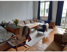 Wohnzimmer mit Holzboden, einer großen L-förmigen Sofalandschaft und einem niedrigen Holztisch in der Mitte. Große Fenster mit blauen Vorhängen lassen viel Tageslicht herein; im Vordergrund ein Sessel mit einer Decke und eine Vase mit Tulpen auf einem Beistelltisch.