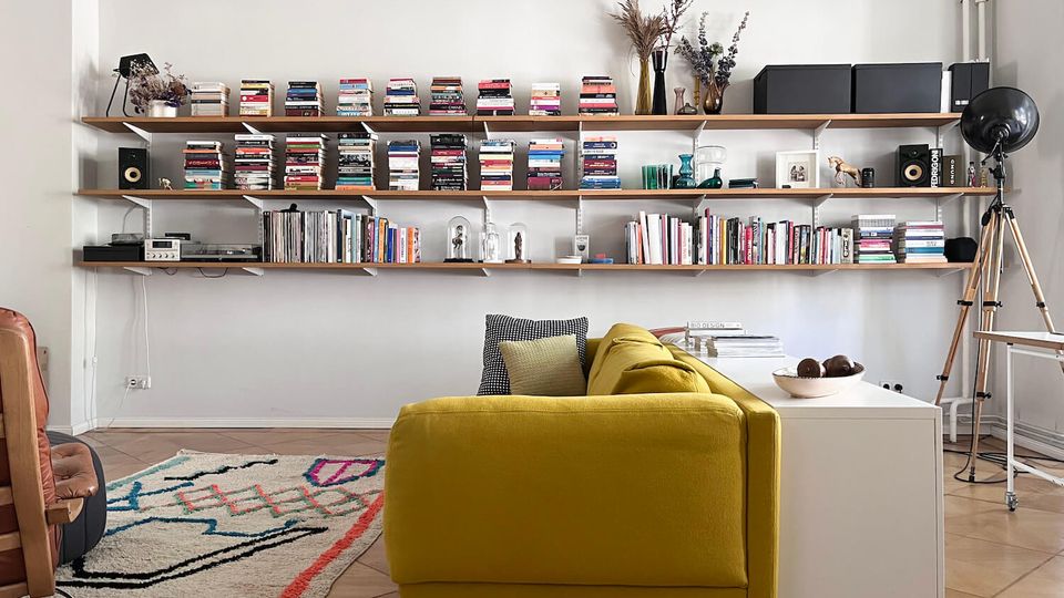 Modern living room with a mustard-yellow sofa facing a long wall of floating wooden shelves filled with books, records, speakers, and decorative vases on a white wall. A patterned area rug and a white side table sit on tan tile flooring, with a large black tripod floor lamp in the corner.