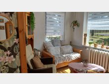 Heller Wohnbereich mit Rattansofa und Sessel, dekoriert mit Kissen, Decken und Pflanzen, neben zwei großen Fenstern mit Doppelrollos. Vor den Fenstern steht ein Heizkörper; auf der Fensterbank befinden sich kleine Dekofiguren und Topfpflanzen, davor ein kleiner Couchtisch.