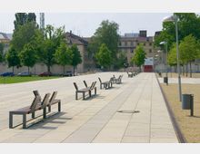 Weitläufiger Platz mit gepflastertem Boden, modern gestalteten Sitzbänken in regelmäßigen Abständen und Bäumen entlang einer Grünfläche. Im Hintergrund sind mehrstöckige Gebäude und einige parkende Autos zu sehen.