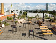 Dachterrasse mit Sitz- und Stehmöglichkeiten, bestehend aus Holzbänken, Tischen und Pflanzenkübeln. Im Hintergrund sind moderne Gebäude, ein hoher Schornstein und eine grüne Umgebung zu sehen.