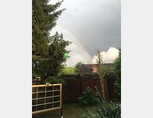 Ein Garten mit hohen Nadelbäumen, Holzzaun und einer kleinen roten Hausfassade im Hintergrund; darüber ein Regenbogen vor einem bewölkten Himmel. Im Vordergrund steht eine Holzrahmenkonstruktion mit Glasplatten.