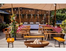 Überdachte Außenterrasse mit Holzboden, langen Bierzeltgarnituren und rot-weiß karierten Tischdecken; darüber hängen Lichterketten und Pflanzenranken. Im Hintergrund steht eine rustikale Holzwand mit Buffetaufbau, seitlich sind Topfpflanzen und ein Grill zu sehen.