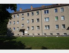 Mehrfamilienhaus mit vier Stockwerken und verputzter Fassade, vielen rechteckigen Fenstern und einem zentralen Eingang. Davor befindet sich eine Rasenfläche, umgeben von Bäumen und Gehwegen.