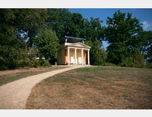 Ein kleines, klassizistisches Gebäude mit Säulenvorhalle steht inmitten eines Parks, umgeben von Bäumen. Ein geschwungener Kiesweg führt durch eine trockene Wiese zu dem Gebäude.