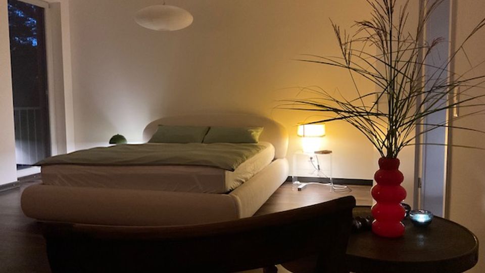 Modernes Schlafzimmer mit niedrigem, gepolstertem Bett und grüner Bettwäsche, Hängelampe sowie Stehlampe neben dem Bett. Große Fensterflächen, dunkler Boden und ein Beistelltisch mit roter Vase und hohen Zweigen im Vordergrund.