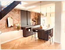 Moderne Küche mit freiliegender Backsteinwand, dunkler Einbauküche, einer Kücheninsel mit dunkler Arbeitsplatte sowie Hockern darunter. Darüber hängen drei goldene Pendelleuchten; daneben ein Spiegel mit Holzrahmen und ein Blumenstrauß auf weißer Ablage.