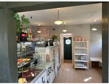 Ein kleines Café mit einer Glasvitrine voller Kuchen und Desserts im Vordergrund, darüber Flaschen mit Getränken. Im Hintergrund eine grüne Tür, eine Preistafel an der Wand und ein Regal mit Produkten wie Milchpackungen und Kaffeebohnen.