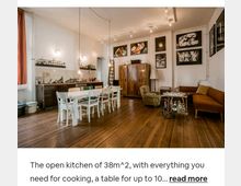 Offene Küche und Wohnraum mit Holzdielenboden, einem großen Esstisch mit Stühlen, minimalistisch gestalteter Küchenzeile an der Wand, und einem gemütlichen Sitzbereich mit Sofa, Sesseln und Barwagen. Wände sind mit gerahmten Bildern dekoriert, und das Ambiente wird durch Hängelampen und natürliche Beleuchtung ergänzt.