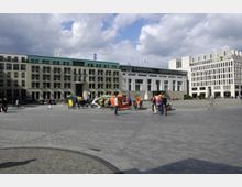 Weitläufiger Platz mit moderner Architektur im Hintergrund, bestehend aus mehrstöckigen Gebäuden mit rechteckigen Fenstern und Glasdächern. Im Vordergrund stehen mehrere Fahrradrikschas in unterschiedlichen Farben, mit kleinen Gruppen von Menschen darum herum.