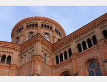 Backsteinfassade einer historischen Kirche mit reich verzierten Bögen, Säulen und Fenstern. Im oberen Bereich ist eine Kuppel mit Rundbogenarkaden und einem Kreuz an der Spitze sichtbar, vor klarem Himmel.