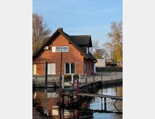 Backsteinbau mit Satteldach am Ufer eines Gewässers, davor ein kleiner Steg mit Pfosten. Im Hintergrund sind Bäume und ein umzäuntes Gelände sichtbar.