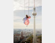 Blick über die Berliner Innenstadt mit dem Fernsehturm am Alexanderplatz im Hintergrund. Im Vordergrund hängt eine Schaukel an Ketten hoch über den Dächern, darunter sind Straßen, Gebäude und Baumreihen zu sehen.