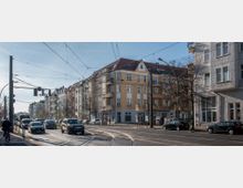 Straßenszene in einer Stadt mit mehrstöckigen Wohngebäuden im Altbaustil, gelber und weißer Fassade sowie roten Dächern. Vor den Gebäuden verlaufen Straßenbahn-Schienen, und die Fahrbahn ist von Autos, einem Radfahrer und Fußgängern frequentiert; Oberleitungen für die Straßenbahn sind sichtbar.